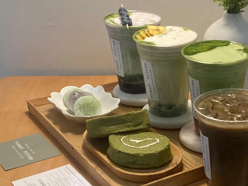 Matcha drinks, matcha roll cake and mochi on a wood tray, with the shop's printed cards on the table